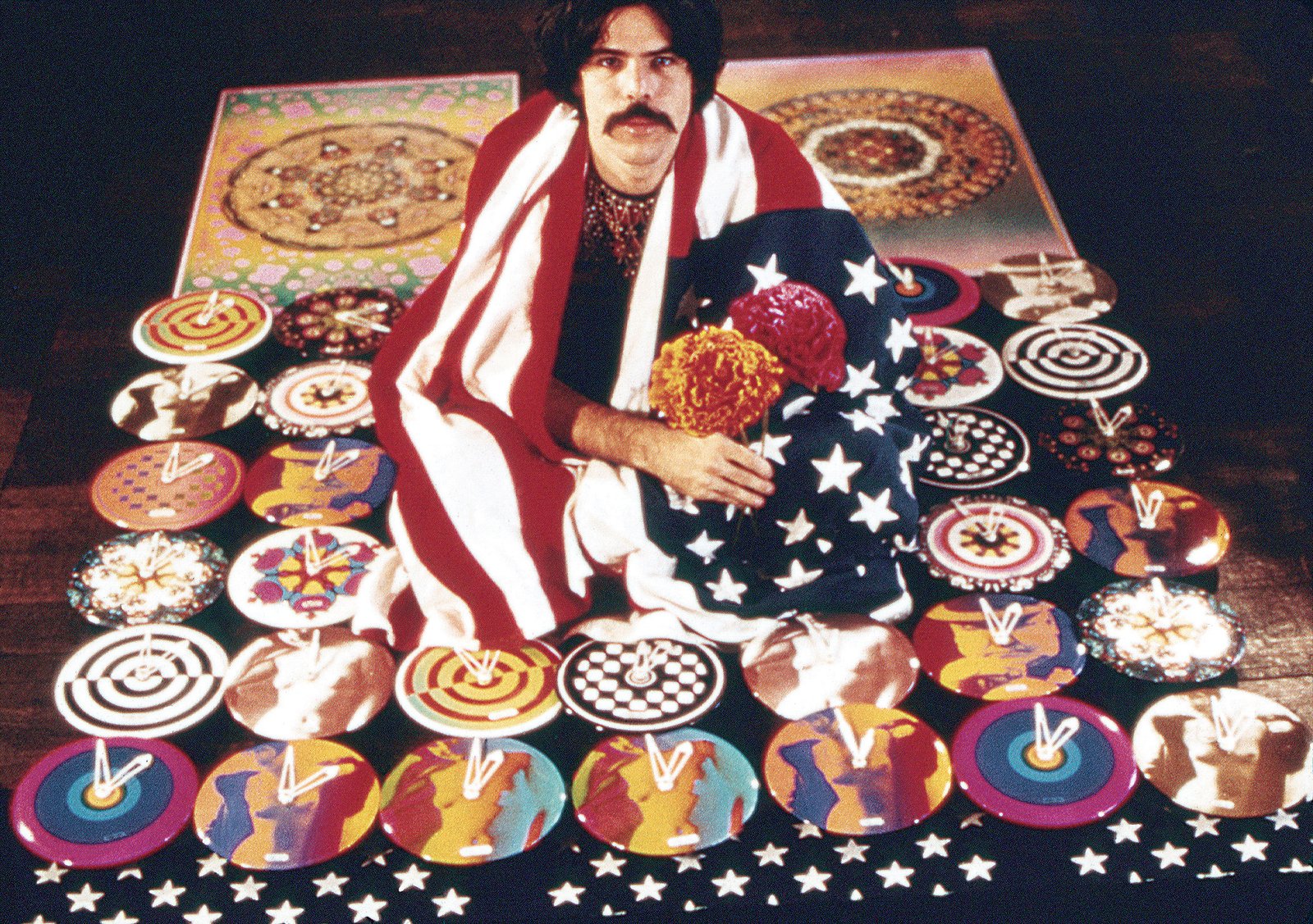 Peter Max wrapped in an American flag crouches over his General Electric art clocks