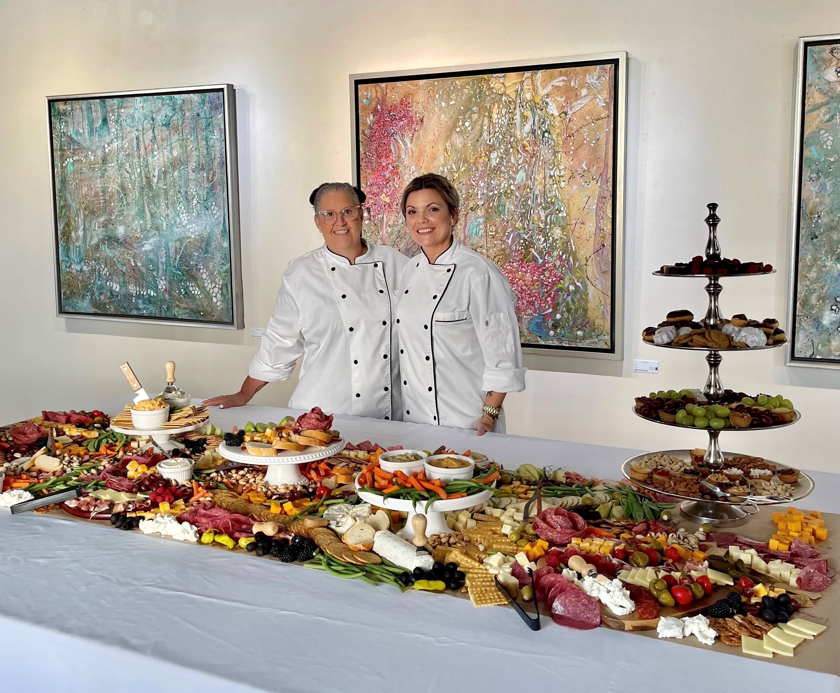 Chefs smile behind a table of charcuterie in front of framed abstract paintings