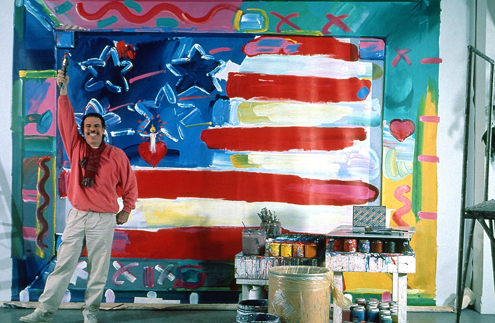 Peter Max holds brush in front of large American flag painting