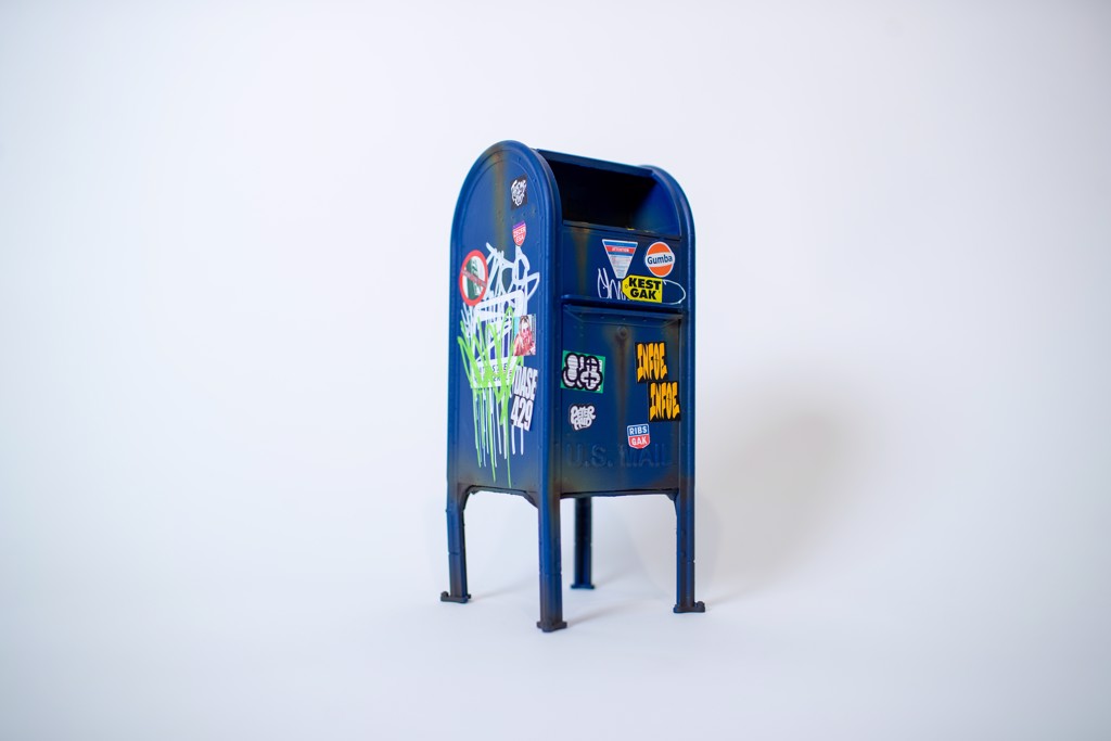 Danny Cortes Mailbox: miniature blue street mailbox on four legs with colorful stickers and graffiti tags.
