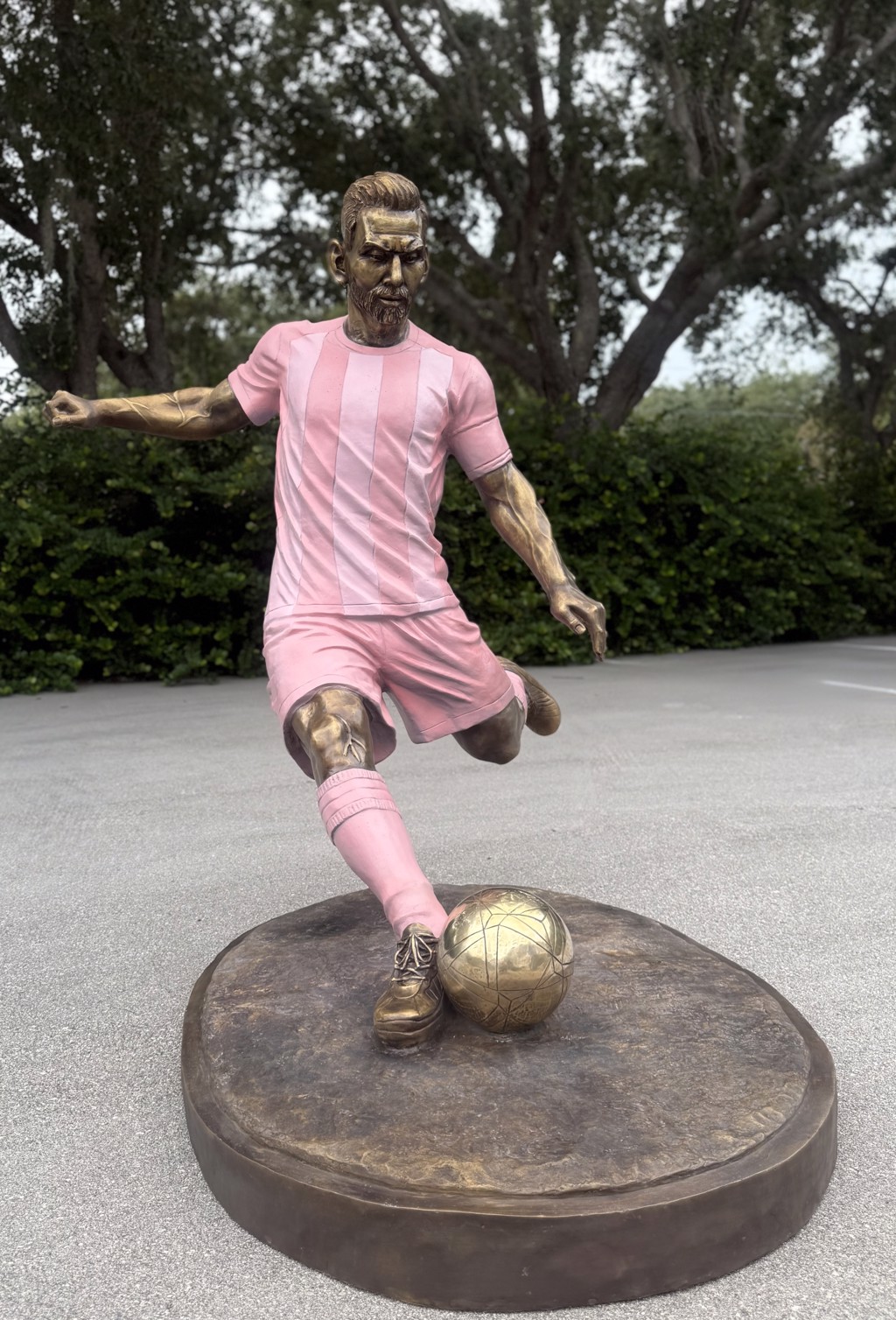 Matthew Snider Leo Messi sculpture: a bronze footballer in a pink uniform prepares to kick a ball on an oval bronze-colored base.