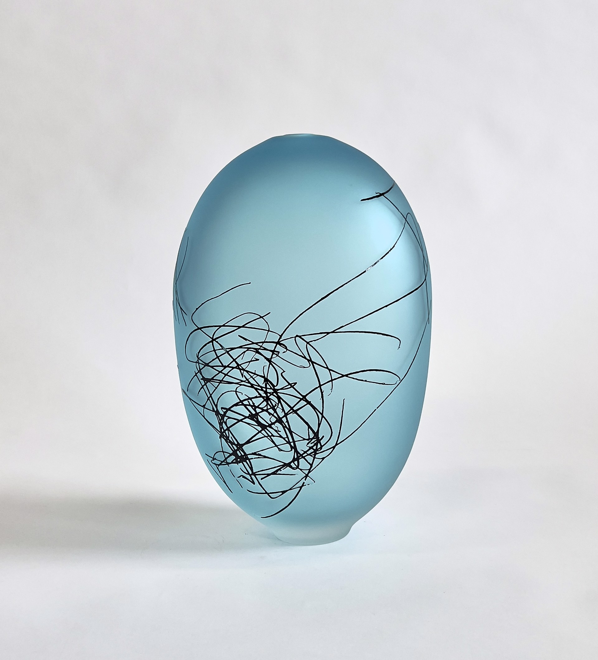 Copper Blue Scribe Ovoid by The Goodman Studios is a striking glass sculpture with a sandblasted surface, featuring intricate markings and a mesmerizing light blue hue. The abstract design invites contemplation and appreciation of the interplay between light, shadow, and form.