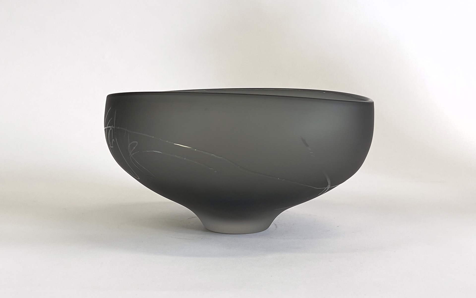 The Goodman Studios Neutral Grey Scribe Bowl is striking glass sculpture with a sandblasted surface, featuring intricate clear markings and a elegant grey hue. The abstract design invites contemplation and appreciation of the interplay between light, shadow, and form.