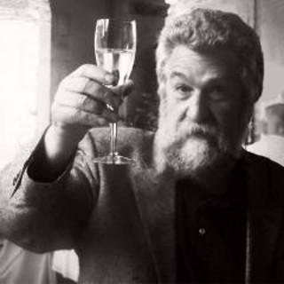 Black and white photograph of fine artist Fred Wessel raising a glass of wine in a toast. Wessel, with a thick beard and wearing a textured blazer over a dark shirt, gazes warmly toward the camera in a celebratory gesture. The soft lighting and vintage feel of the image lend a timeless, inviting quality, capturing a moment of camaraderie and appreciation. Black and white photograph of fine artist Fred Wessel raising a glass of wine in a toast. Wessel, with a thick beard and wearing a textured blazer over a dark shirt, gazes warmly toward the camera in a celebratory gesture. The soft lighting and vintage feel of the image lend a timeless, inviting quality, capturing a moment of camaraderie and appreciation.