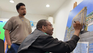 An artist wearing glasses, seated and working on a detailed architectural painting of a cityscape, as another individual stands nearby observing the process in a studio environment An artist wearing glasses, seated and working on a detailed architectural painting of a cityscape, as another individual stands nearby observing the process in a studio environment