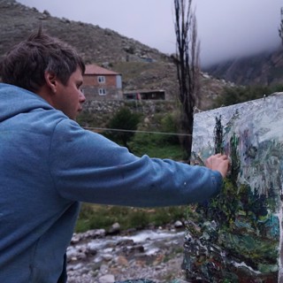Fine artist Ulrich Gleiter painting en plein air in a mountainous landscape, likely in Germany. Dressed in a blue hoodie, Gleiter focuses intensely on his canvas, applying textured strokes with his hands, a hallmark of his expressive and impressionistic style. The painting depicts a rugged natural scene with lush greens and muted earth tones, mirroring the surrounding environment of rocky hills, scattered trees, and a nearby stream. Mist and fog drift over the distant mountains, enhancing the atmosphere and sense of immersion in nature. This scene captures Gleiter’s dedication to capturing landscapes directly from life with dynamic, tactile brushwork. Fine artist Ulrich Gleiter painting en plein air in a mountainous landscape, likely in Germany. Dressed in a blue hoodie, Gleiter focuses intensely on his canvas, applying textured strokes with his hands, a hallmark of his expressive and impressionistic style. The painting depicts a rugged natural scene with lush greens and muted earth tones, mirroring the surrounding environment of rocky hills, scattered trees, and a nearby stream. Mist and fog drift over the distant mountains, enhancing the atmosphere and sense of immersion in nature. This scene captures Gleiter’s dedication to capturing landscapes directly from life with dynamic, tactile brushwork.