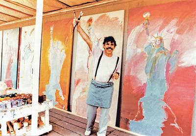 Peter Max smiling holding brush in front of a series of Lady Liberty paintings